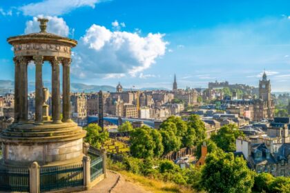 View of Edinburgh from Calton Hill on a sunny day, showing the Dugald Stewart Monument in the foreground and a panoramic skyline featuring Edinburgh Castle, the Balmoral Hotel clock tower, and the spires of the Old Town, with the Pentland Hills in the background.