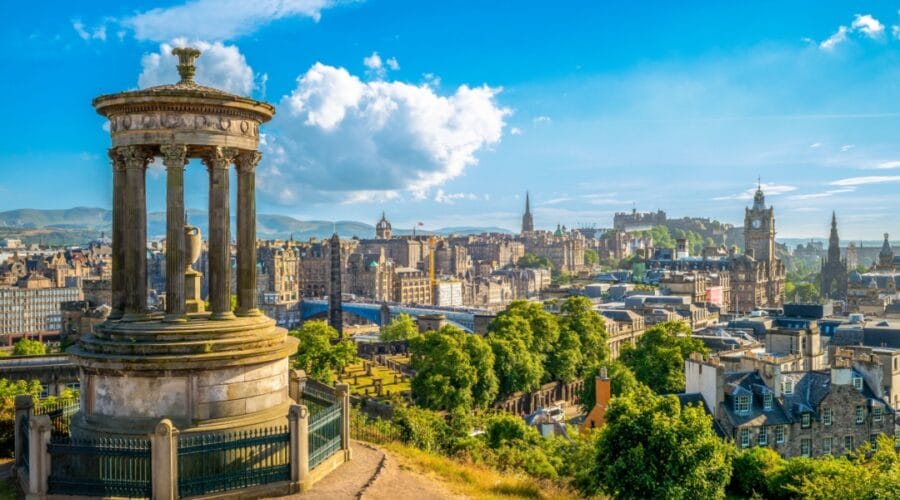 View of Edinburgh from Calton Hill on a sunny day, showing the Dugald Stewart Monument in the foreground and a panoramic skyline featuring Edinburgh Castle, the Balmoral Hotel clock tower, and the spires of the Old Town, with the Pentland Hills in the background.