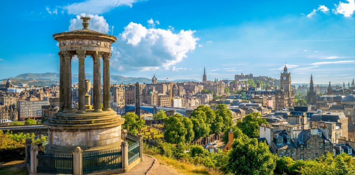 View of Edinburgh from Calton Hill on a sunny day, showing the Dugald Stewart Monument in the foreground and a panoramic skyline featuring Edinburgh Castle, the Balmoral Hotel clock tower, and the spires of the Old Town, with the Pentland Hills in the background.