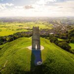 Aerial view of Glastonbury Tor in Somerset, with the medieval St Michael’s Tower standing atop a lush green hill, overlooking a patchwork of fields and countryside under a partly cloudy sky