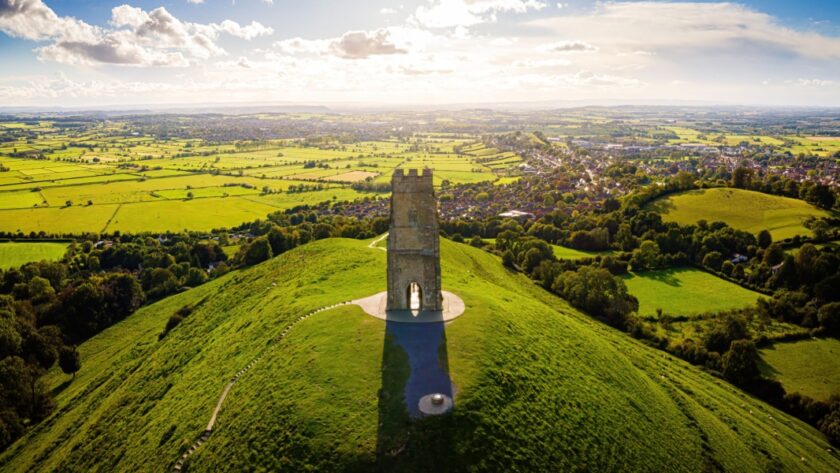 Aerial view of Glastonbury Tor in Somerset, with the medieval St Michael’s Tower standing atop a lush green hill, overlooking a patchwork of fields and countryside under a partly cloudy sky