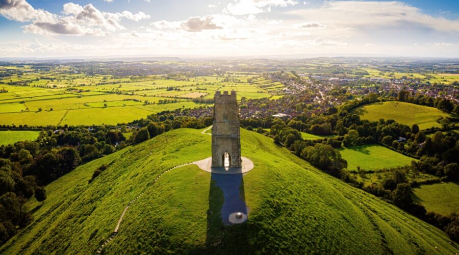 Aerial view of Glastonbury Tor in Somerset, with the medieval St Michael’s Tower standing atop a lush green hill, overlooking a patchwork of fields and countryside under a partly cloudy sky