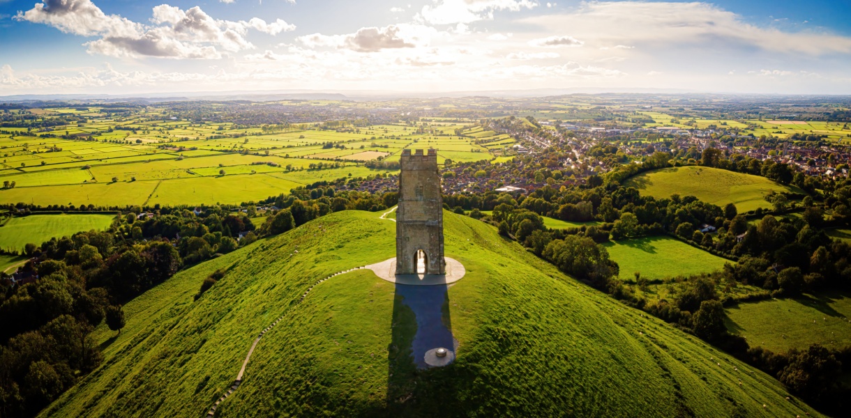 Aerial view of Glastonbury Tor in Somerset, with the medieval St Michael’s Tower standing atop a lush green hill, overlooking a patchwork of fields and countryside under a partly cloudy sky