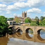 View of Hereford in Herefordshire, featuring the historic stone bridge over the River Wye with Hereford Cathedral in the background on a sunny day with blue skies and scattered clouds.