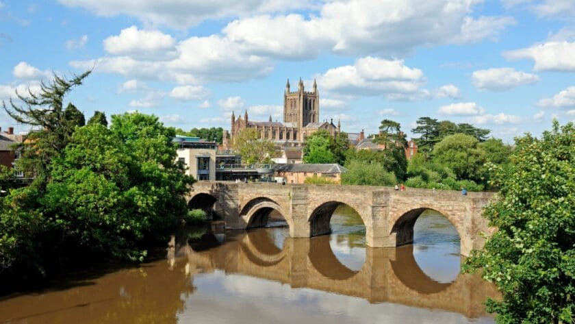 View of Hereford in Herefordshire, featuring the historic stone bridge over the River Wye with Hereford Cathedral in the background on a sunny day with blue skies and scattered clouds.