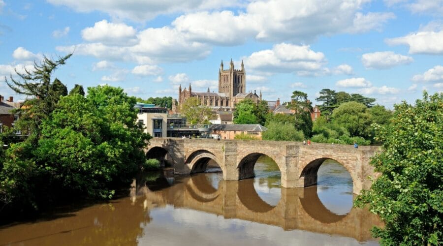 View of Hereford in Herefordshire, featuring the historic stone bridge over the River Wye with Hereford Cathedral in the background on a sunny day with blue skies and scattered clouds.