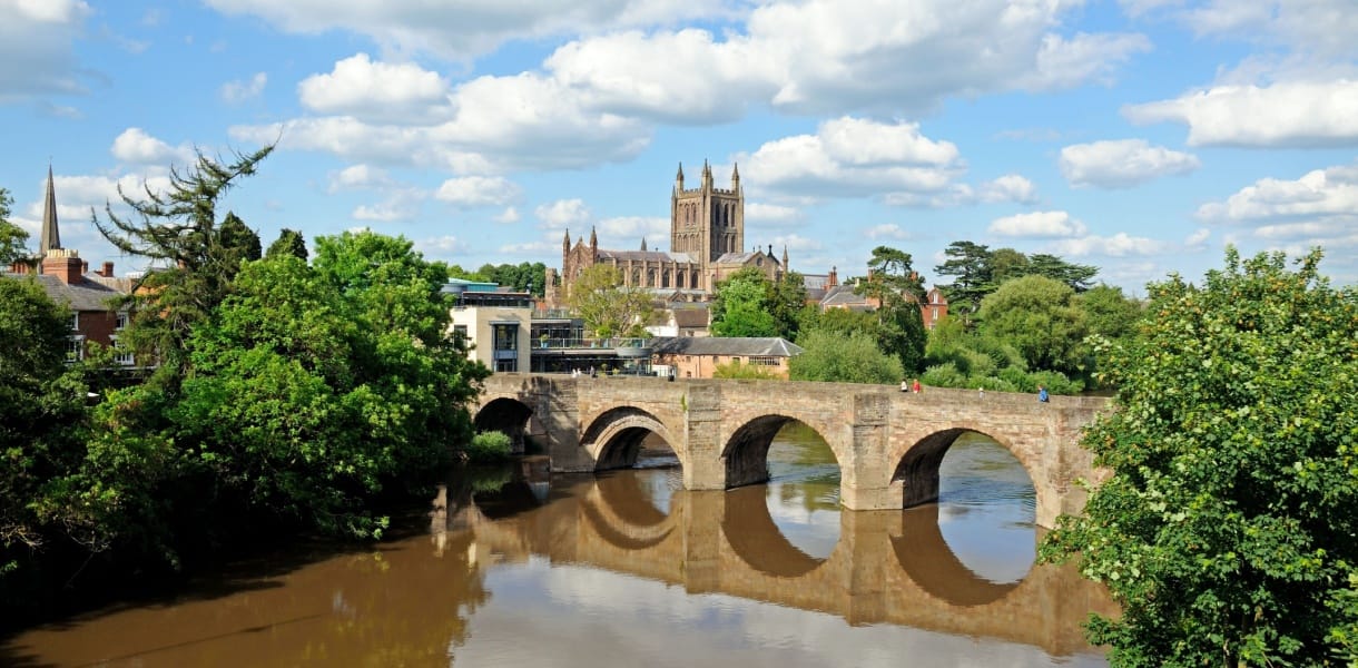View of Hereford in Herefordshire, featuring the historic stone bridge over the River Wye with Hereford Cathedral in the background on a sunny day with blue skies and scattered clouds.
