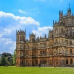 Highclere Castle in Hampshire on a sunny day, with its grand Victorian architecture, ornate towers, and expansive green lawn surrounded by trees under a bright blue sky with scattered clouds