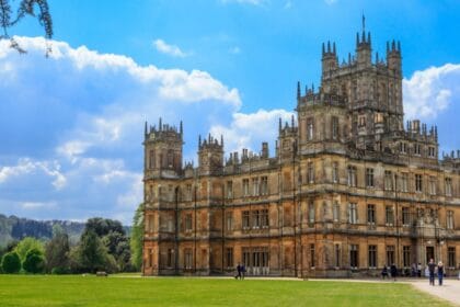Highclere Castle in Hampshire on a sunny day, with its grand Victorian architecture, ornate towers, and expansive green lawn surrounded by trees under a bright blue sky with scattered clouds