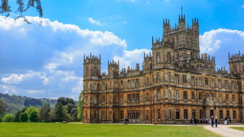 Highclere Castle in Hampshire on a sunny day, with its grand Victorian architecture, ornate towers, and expansive green lawn surrounded by trees under a bright blue sky with scattered clouds