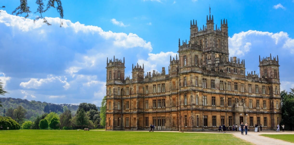 Highclere Castle in Hampshire on a sunny day, with its grand Victorian architecture, ornate towers, and expansive green lawn surrounded by trees under a bright blue sky with scattered clouds