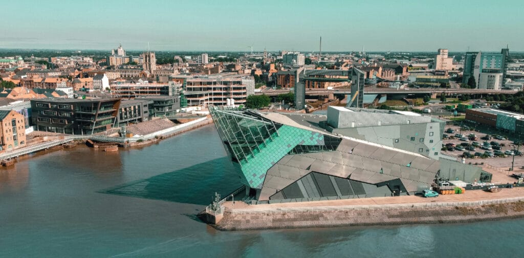 Aerial view of The Deep aquarium in Hull, East Yorkshire, with its distinctive angular glass-and-steel architecture jutting out over the River Humber. The surrounding cityscape features a mix of historic buildings, modern developments, and the Humber waterfront under a clear blue sky.