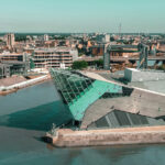 Aerial view of The Deep aquarium in Hull, East Yorkshire, with its distinctive angular glass-and-steel architecture jutting out over the River Humber. The surrounding cityscape features a mix of historic buildings, modern developments, and the Humber waterfront under a clear blue sky.