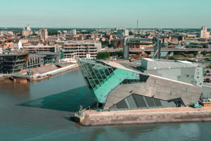 Aerial view of The Deep aquarium in Hull, East Yorkshire, with its distinctive angular glass-and-steel architecture jutting out over the River Humber. The surrounding cityscape features a mix of historic buildings, modern developments, and the Humber waterfront under a clear blue sky.