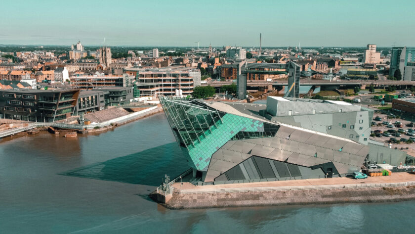 Aerial view of The Deep aquarium in Hull, East Yorkshire, with its distinctive angular glass-and-steel architecture jutting out over the River Humber. The surrounding cityscape features a mix of historic buildings, modern developments, and the Humber waterfront under a clear blue sky.