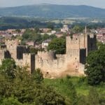 Ludlow Castle in Shropshire, England, viewed from a hillside with lush greenery in the foreground and the historic market town of Ludlow and rolling Shropshire Hills in the background.