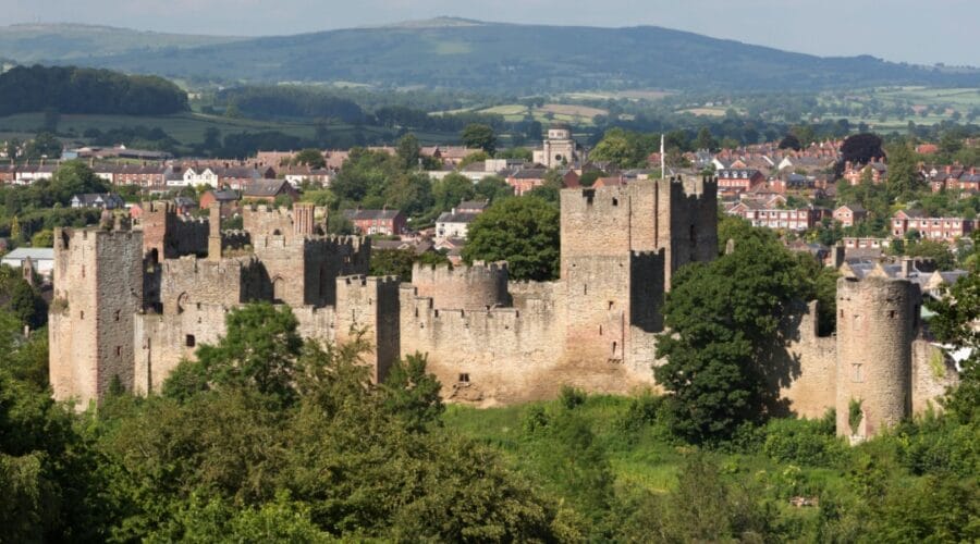 Ludlow Castle in Shropshire, England, viewed from a hillside with lush greenery in the foreground and the historic market town of Ludlow and rolling Shropshire Hills in the background.