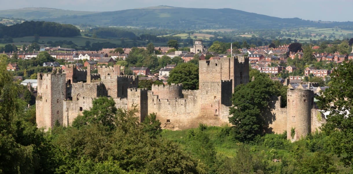 Ludlow Castle in Shropshire, England, viewed from a hillside with lush greenery in the foreground and the historic market town of Ludlow and rolling Shropshire Hills in the background.