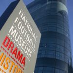 Sign for the National Football Museum in Manchester featuring the words "Drama", "History", and "Skill" in bold colours, with the museum’s modern glass-fronted building rising behind it against a clear blue sky