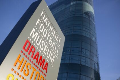 Sign for the National Football Museum in Manchester featuring the words "Drama", "History", and "Skill" in bold colours, with the museum’s modern glass-fronted building rising behind it against a clear blue sky
