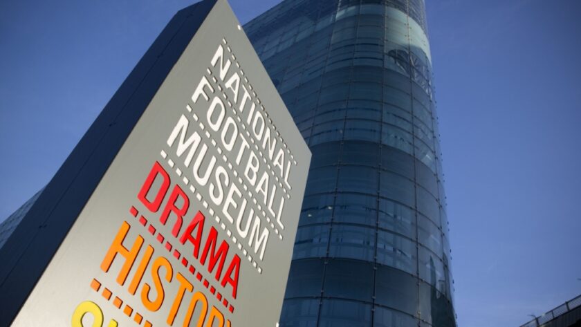 Sign for the National Football Museum in Manchester featuring the words "Drama", "History", and "Skill" in bold colours, with the museum’s modern glass-fronted building rising behind it against a clear blue sky