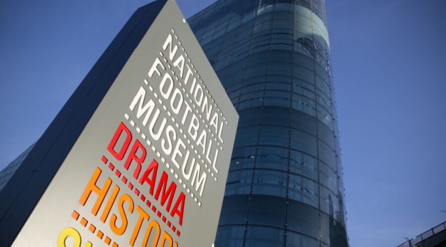 Sign for the National Football Museum in Manchester featuring the words "Drama", "History", and "Skill" in bold colours, with the museum’s modern glass-fronted building rising behind it against a clear blue sky