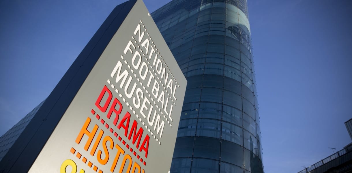 Sign for the National Football Museum in Manchester featuring the words "Drama", "History", and "Skill" in bold colours, with the museum’s modern glass-fronted building rising behind it against a clear blue sky