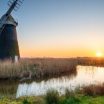 Sunset over Mutton’s Drainage Mill at Halvergate on the Berney Marshes in the Norfolk Broads, with the windmill reflected in a calm water channel surrounded by reeds and grassy marshland.