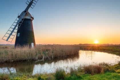 Sunset over Mutton’s Drainage Mill at Halvergate on the Berney Marshes in the Norfolk Broads, with the windmill reflected in a calm water channel surrounded by reeds and grassy marshland.