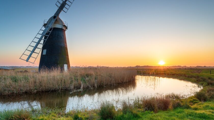 Sunset over Mutton’s Drainage Mill at Halvergate on the Berney Marshes in the Norfolk Broads, with the windmill reflected in a calm water channel surrounded by reeds and grassy marshland.