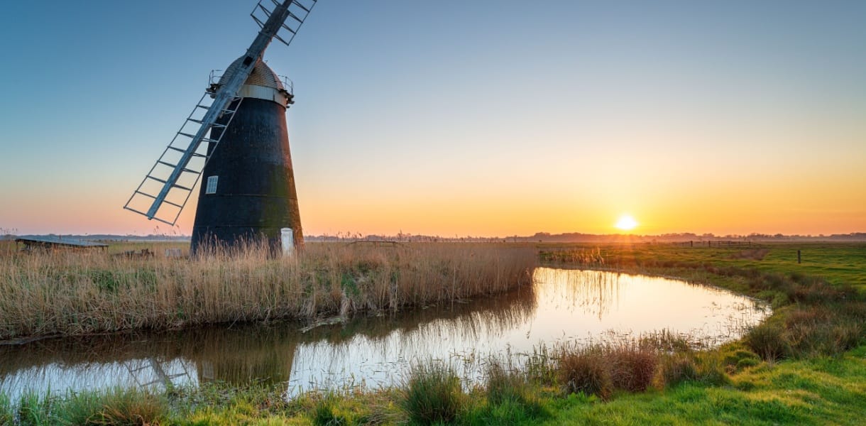 Sunset over Mutton’s Drainage Mill at Halvergate on the Berney Marshes in the Norfolk Broads, with the windmill reflected in a calm water channel surrounded by reeds and grassy marshland.