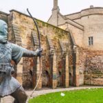 Bronze statue of Robin Hood aiming a bow and arrow in front of the historic stone walls and gatehouse of Nottingham Castle, with red-brick buildings in the background on a cloudy day.