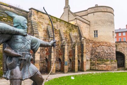 Bronze statue of Robin Hood aiming a bow and arrow in front of the historic stone walls and gatehouse of Nottingham Castle, with red-brick buildings in the background on a cloudy day.