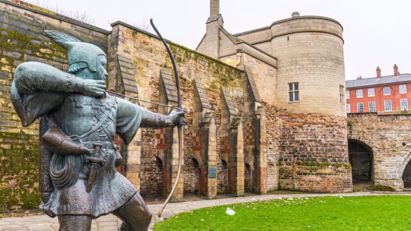 Bronze statue of Robin Hood aiming a bow and arrow in front of the historic stone walls and gatehouse of Nottingham Castle, with red-brick buildings in the background on a cloudy day.