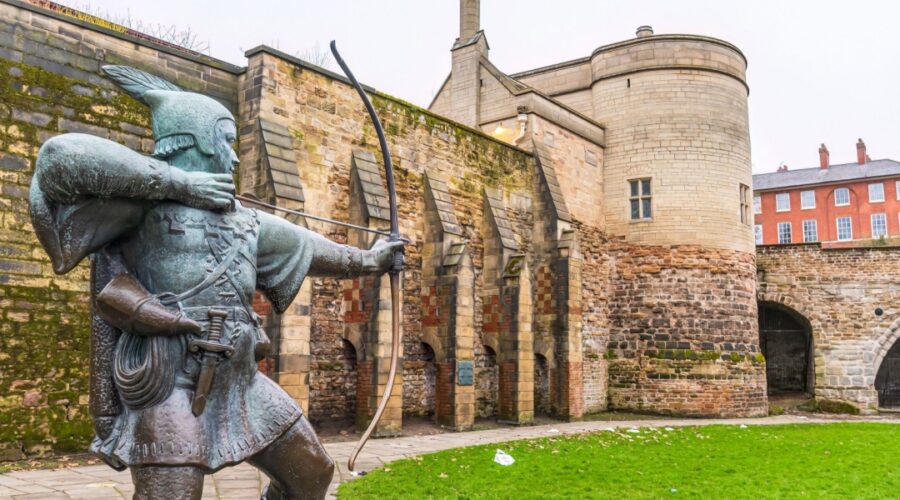 Bronze statue of Robin Hood aiming a bow and arrow in front of the historic stone walls and gatehouse of Nottingham Castle, with red-brick buildings in the background on a cloudy day.