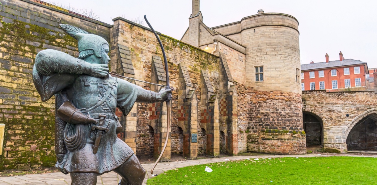 Bronze statue of Robin Hood aiming a bow and arrow in front of the historic stone walls and gatehouse of Nottingham Castle, with red-brick buildings in the background on a cloudy day.