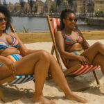 Two young women in colourful bikinis sit on striped deck chairs under a navy beach umbrella on a sandy urban beach at London's Royal Docks. They are smiling and wearing sunglasses, enjoying a sunny day with views of dockside cranes, modern buildings, and calm water in the background.