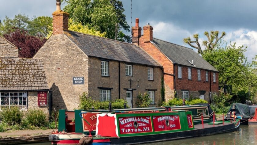 Traditional canal boats moored along the Grand Union Canal in Stoke Bruerne, Northamptonshire, with historic stone cottages and the Canal Shop in the background, surrounded by greenery on a partly cloudy day.