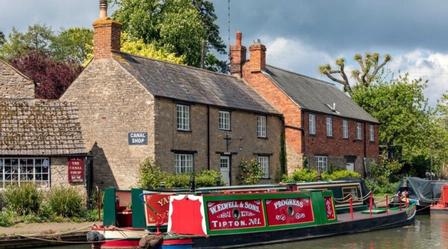 Traditional canal boats moored along the Grand Union Canal in Stoke Bruerne, Northamptonshire, with historic stone cottages and the Canal Shop in the background, surrounded by greenery on a partly cloudy day.
