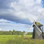 Traditional wooden windmill in a green Suffolk meadow under a dramatic sky with sunlit clouds and blue patches, showcasing the rural charm of the English countryside.