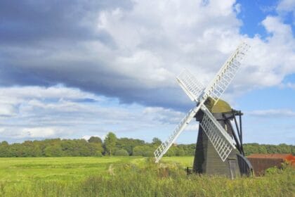 Traditional wooden windmill in a green Suffolk meadow under a dramatic sky with sunlit clouds and blue patches, showcasing the rural charm of the English countryside.