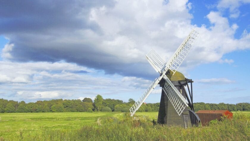 Traditional wooden windmill in a green Suffolk meadow under a dramatic sky with sunlit clouds and blue patches, showcasing the rural charm of the English countryside.