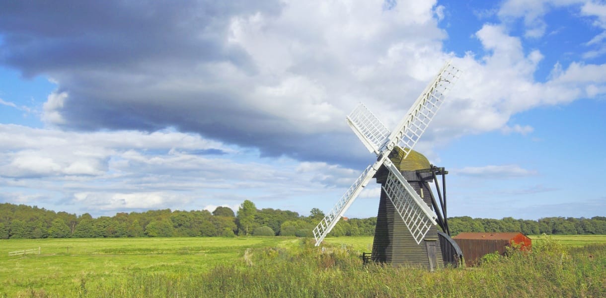 Traditional wooden windmill in a green Suffolk meadow under a dramatic sky with sunlit clouds and blue patches, showcasing the rural charm of the English countryside.