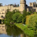 View of Warwick Castle, in Warwickshire, rising above the River Avon, surrounded by lush green trees and reflected in the calm water below, with clear blue skies in the background