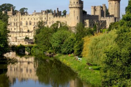 View of Warwick Castle, in Warwickshire, rising above the River Avon, surrounded by lush green trees and reflected in the calm water below, with clear blue skies in the background