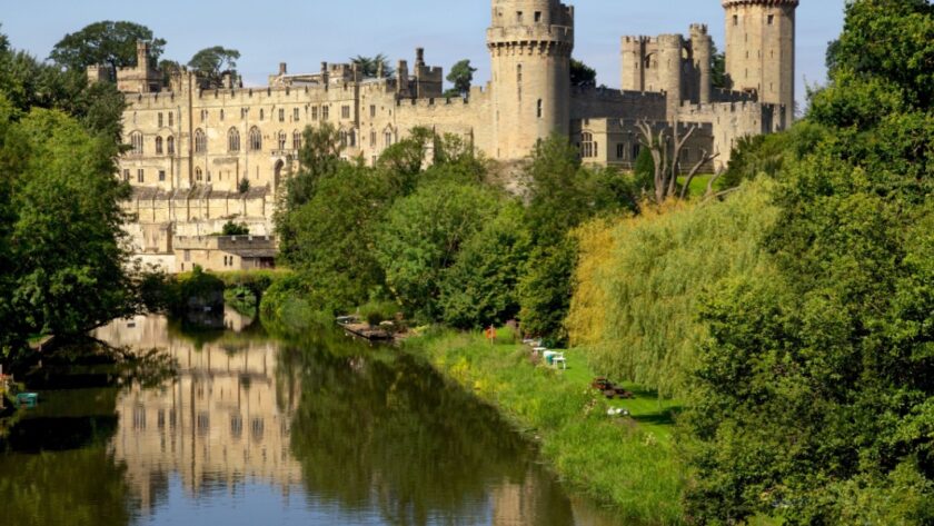 View of Warwick Castle, in Warwickshire, rising above the River Avon, surrounded by lush green trees and reflected in the calm water below, with clear blue skies in the background