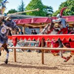 Two knights in full armour jousting on horseback at Warwick Castle, watched by a crowd behind red and gold banners on a sunny day.