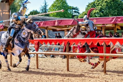 Two knights in full armour jousting on horseback at Warwick Castle, watched by a crowd behind red and gold banners on a sunny day.
