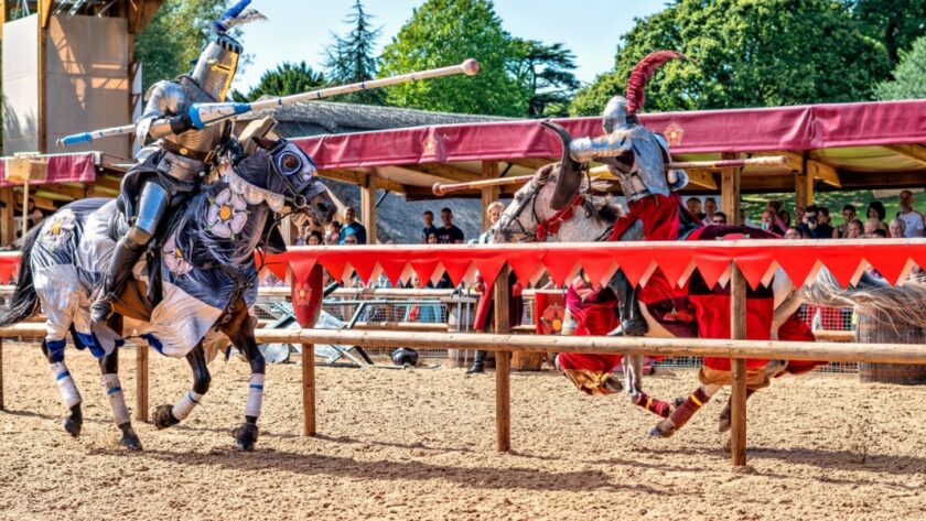 Two knights in full armour jousting on horseback at Warwick Castle, watched by a crowd behind red and gold banners on a sunny day.