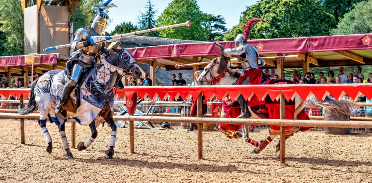 Two knights in full armour jousting on horseback at Warwick Castle, watched by a crowd behind red and gold banners on a sunny day.